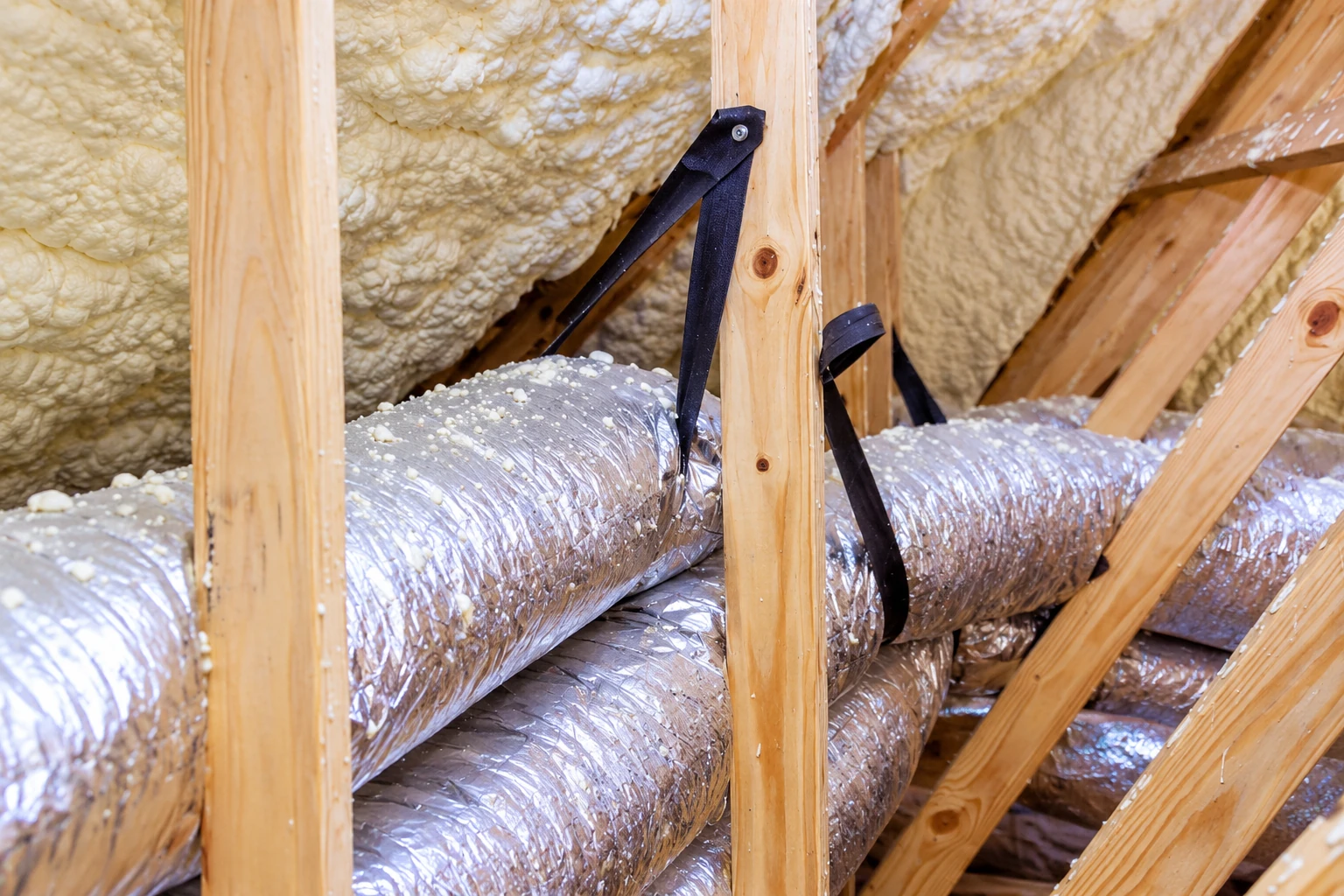 Foam insulation coats roof framing above silver flexible ducts secured with black straps.