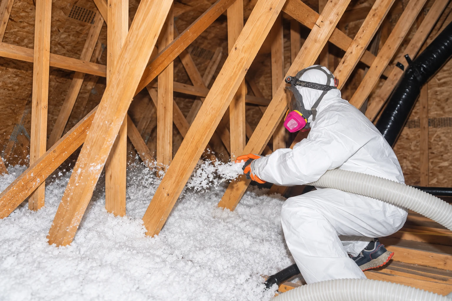 A worker in protective gear uses a hose to blow loose insulation across an attic floor between wooden rafters.