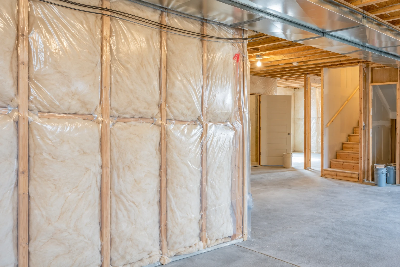 White fiberglass insulation fills plastic-covered basement wall bays beneath exposed ceiling joists.