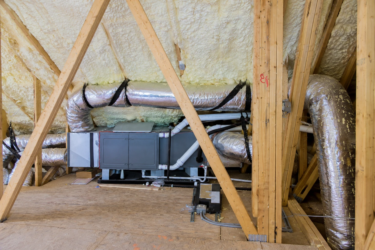 Closed-cell foam covers attic roof framing around silver HVAC ducts and a central air-handling unit.