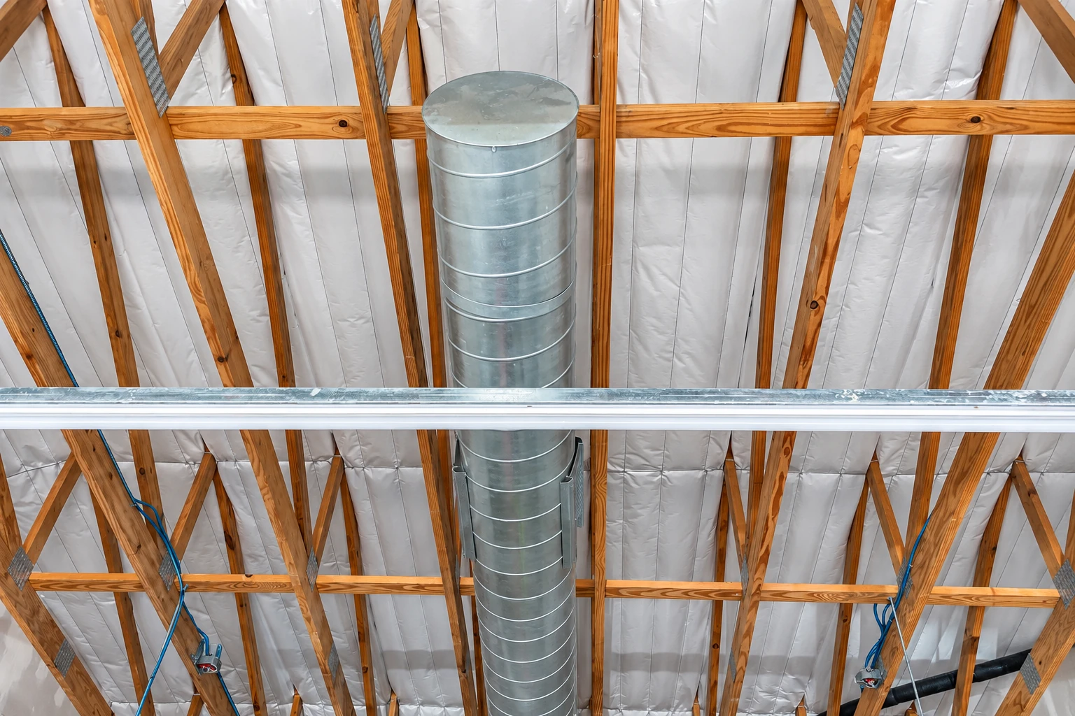 Commercial insulation lines a timber-truss ceiling around a large galvanized ventilation duct.