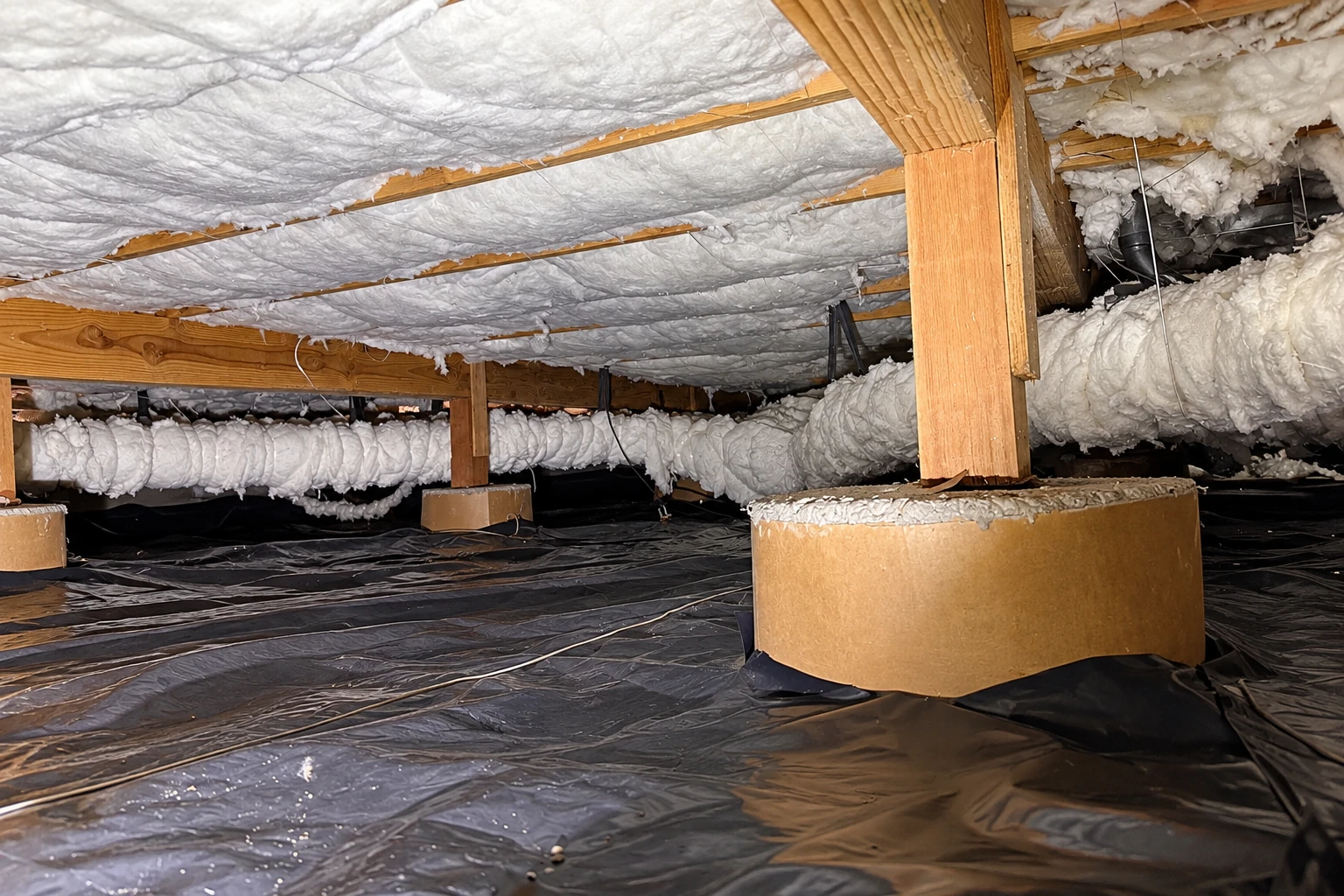 A crawl space has white insulation beneath floor joists, insulated ducts, support piers and a black plastic ground barrier.