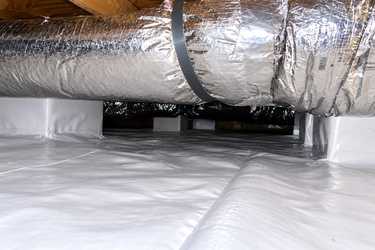 A crawl space is covered with white vapor barrier beneath a wide foil-wrapped duct.