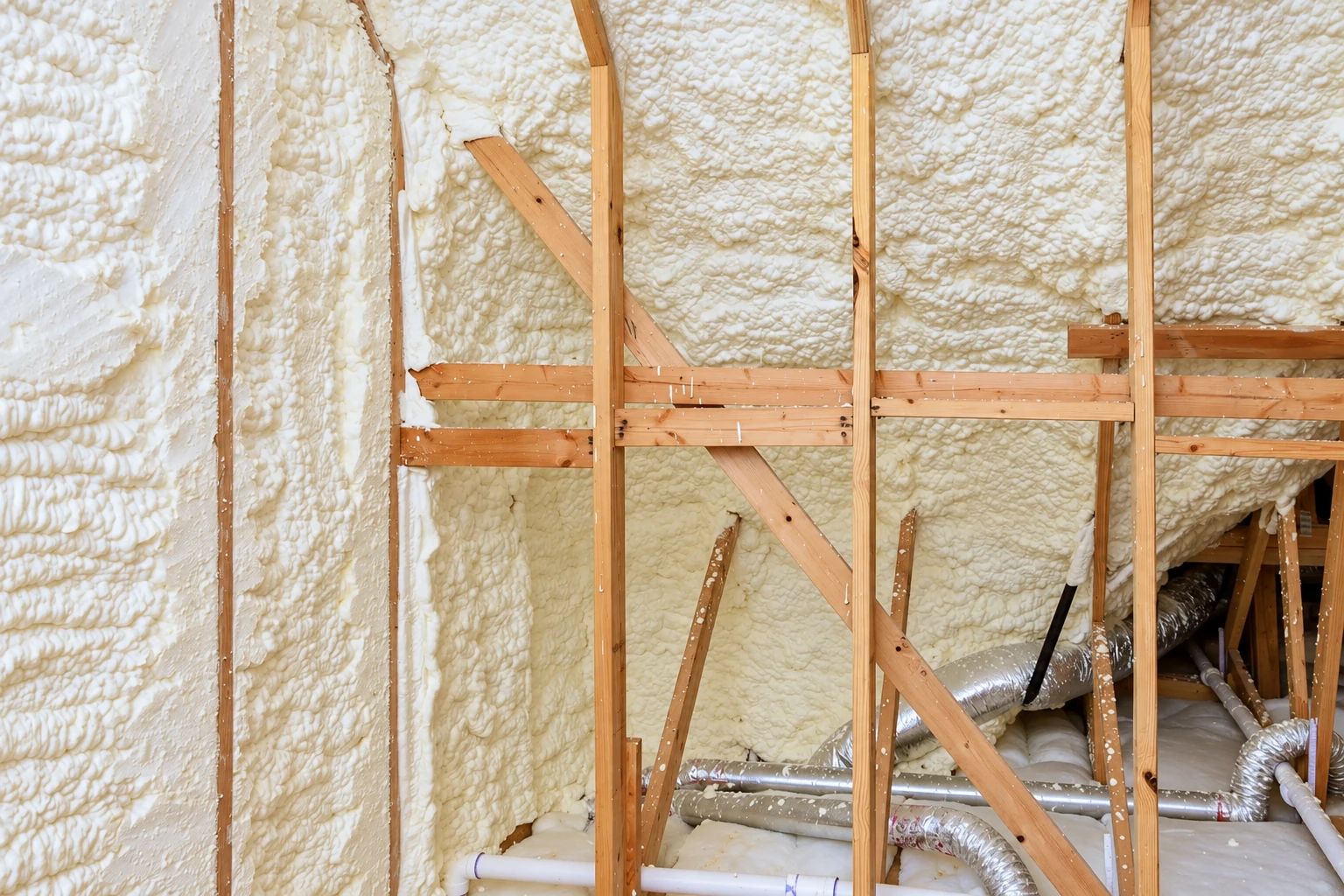 Thick white spray foam covers the walls and roof of a wooden-framed attic with exposed beams and ducts.