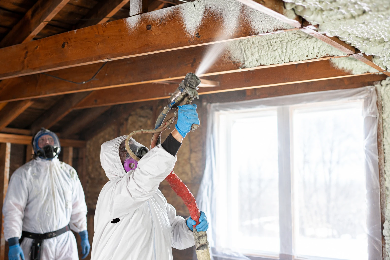 A worker in protective coveralls sprays foam insulation onto exposed roof rafters while another stands nearby.